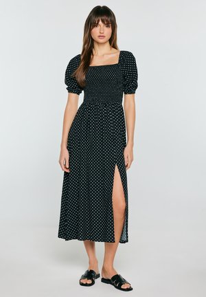 Kobieta w czarnej sukience midi w białe grochy, z bufiastymi rękawami, kwadratowym dekoltem, rozcięciem po boku i czarnych sandałach na płaskiej podeszwie, stojąca na szarym tle.