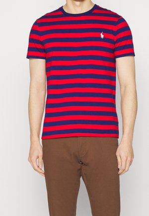 Homme portant un t-shirt à manches courtes rayé horizontalement rouge et bleu marine avec un pantalon marron, debout sur un fond uni.