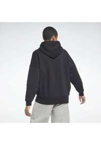 Schwarzer Kapuzenpullover mit lässigem Schnitt, der eine weiche Textur und gerippte Manschetten aufweist. Getragen mit hellgrauen Jogginghosen.
