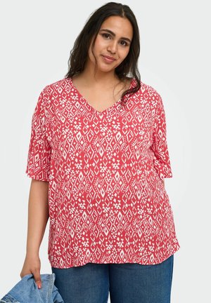 Woman with long dark hair wearing a red and white patterned V-neck shirt and blue jeans, holding a denim jacket in one hand.