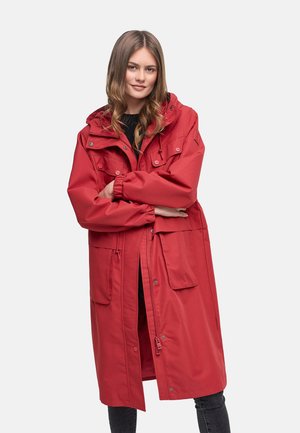 Noor naine pikkade juustega, kes kannab pikka punast kapuutsiga mantlit suurte taskutega, seisab käed rüpes valgel taustal.