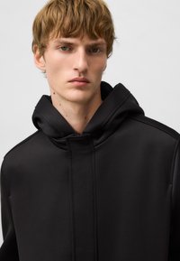 Veste noire à capuche avec une texture lisse, dotée d'un zip caché, de coutures détaillées et d'une coupe décontractée au niveau du col.