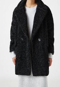 Manteau noir texturé à double boutonnage avec un large col et deux boutons. Porté par-dessus un pull gris clair côtelé. Matière douce et pelucheuse.