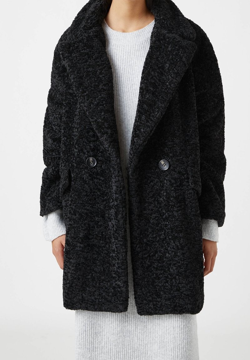 Manteau noir texturé à double boutonnage avec un large col et deux boutons. Porté par-dessus un pull gris clair côtelé. Matière douce et pelucheuse.