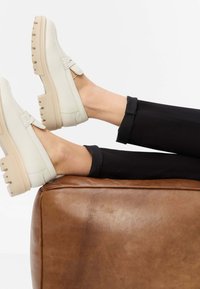 Witte leren schoenen met een gestructureerd oppervlak en beige stevige zolen, geplaatst op een bruine leren kussen, gecombineerd met zwarte opgerolde broekspijpen.