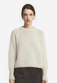 Beige stickad tröja med rund halsringning, ribbad mudd och fåll. Har en avslappnad passform och texturerad tyg, kombinerad med en mörk kjol.