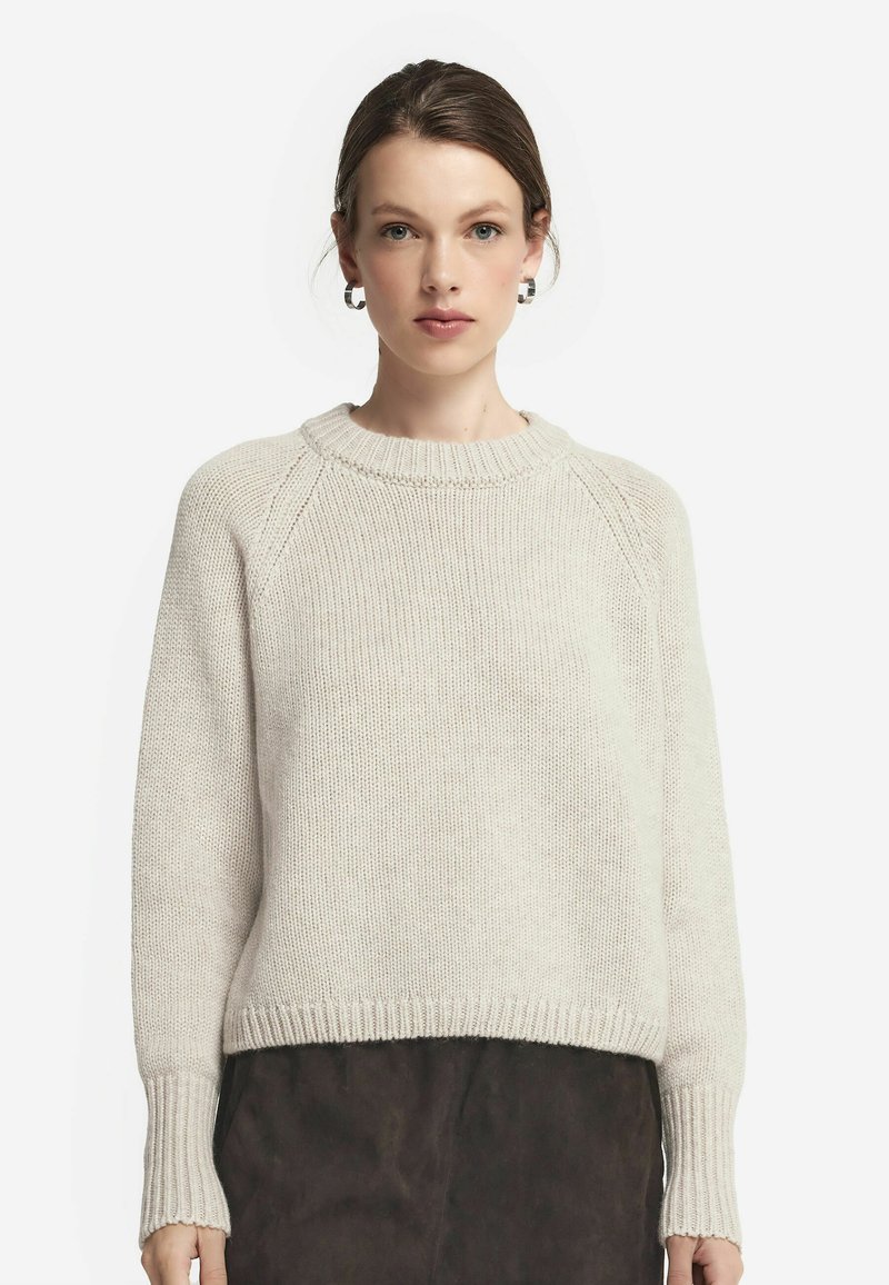 Beige stickad tröja med rund halsringning, ribbad mudd och fåll. Har en avslappnad passform och texturerad tyg, kombinerad med en mörk kjol.