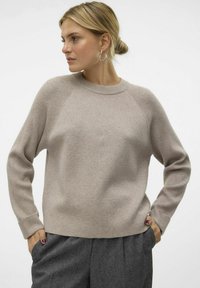 Beige gestrickter Pullover mit geripptem Hals- und Bündchen, mit Raglanärmeln und lockerer Passform, kombiniert mit grauen strukturierten Hosen.
