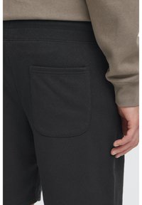 Un short noir en tissu doux comporte une poche arrière avec un bord supérieur arrondi. Fini avec une ceinture côtelée. Le tissu présente une légère texture.