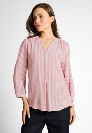 Frau trägt eine rosa-weiß gestreifte Bluse mit vertikalen Streifen, drei Knöpfen und dreiviertellangen Ärmeln, kombiniert mit dunkler Hose.