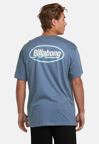 Blå bomulds T-shirt med et stort hvidt og lyseblåt "Billabong" logo på ryggen. Korte ærmer og en afslappet pasform.