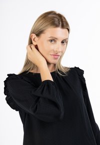 Blusa nera in tessuto texture, maniche a sbuffo e dettagli arricciati. La modella ha i capelli biondi e uno sfondo neutro.