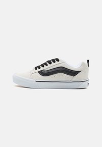 Adidași cu un superior din suede gri deschis, accente din piele neagră, talpă intermediară albă și șireturi negre. Prezintă perforații și un logo pe toc.
