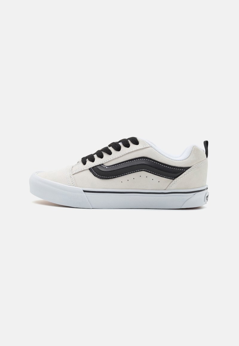 Adidași cu un superior din suede gri deschis, accente din piele neagră, talpă intermediară albă și șireturi negre. Prezintă perforații și un logo pe toc.