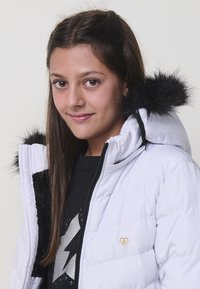 Chaqueta acolchada blanca con capucha de piel sintética negra, que presenta un emblema de corazón dorado, diseño acolchado y cremallera lateral.