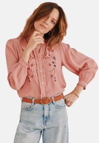 Rosa Bluse mit floralem Stickerei, gerafftem Vorderteil und Knopfverschluss. Verfügt über lange Ärmel und eine lockere Passform, kombiniert mit hellblauen Jeans.