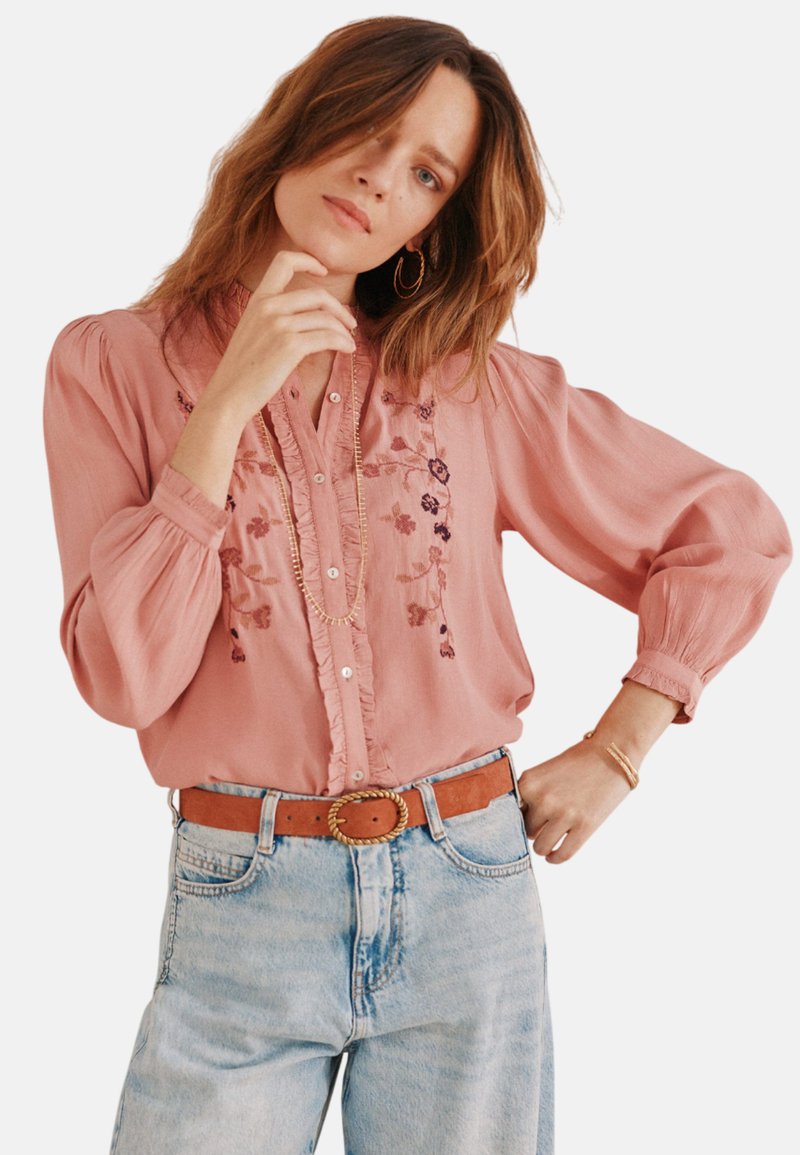 Rosa Bluse mit floralem Stickerei, gerafftem Vorderteil und Knopfverschluss. Verfügt über lange Ärmel und eine lockere Passform, kombiniert mit hellblauen Jeans.