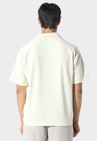 Krátký rukáv košile z off-white vaflové pleteniny; má špičatý límec a jemné švy na zadní části.