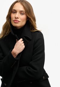 Zwarte wollen jas met hoge kraag, dubbele rij met knopen en zijzakken. Warme textuur met een getailleerd silhouet.