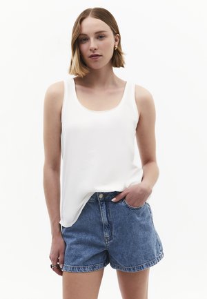 Donna con capelli corti biondi che indossa una canotta bianca senza maniche e pantaloncini di jeans blu, in piedi con una mano in tasca su sfondo bianco.