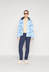 Light blue patterned jacket with a zip front and hood, paired with navy pants and beige shoes. Model wearing sunglasses.