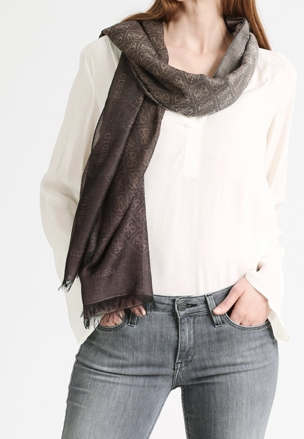 Femme portant une écharpe marron foncé à motifs sur un chemisier blanc et un jean gris, avec une main dans la poche, sur un fond uni.