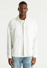 Witte blouse met een relaxte pasvorm, lange mouwen, borstzakken, gedragen over een lichtgrijs T-shirt, gecombineerd met lichtblauwe spijkerbroek.