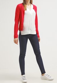 Cardigan rouge, blouse blanche et jean foncé. Le cardigan se ferme par des boutons, et la blouse présente un col en V avec un bord festonné. Des baskets blanches complètent le look.