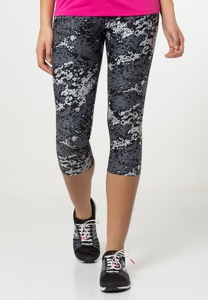 Frau trägt schwarze und graue, pixelartige Leggings und schwarze Laufschuhe und geht vorwärts auf weißem Hintergrund.