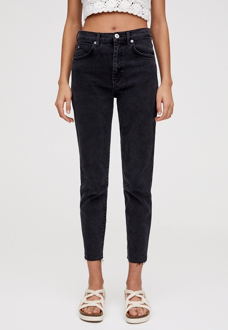 Femme portant un jean noir slim taille haute, un crop top en crochet blanc et des sandales blanches en corde, debout devant un fond clair uni.