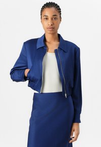Giacca bomber cropped navy in tessuto morbido, con colletto a punta, chiusura con zip e dettaglio di orlo arricciato. Indossata sopra una maglietta chiara.