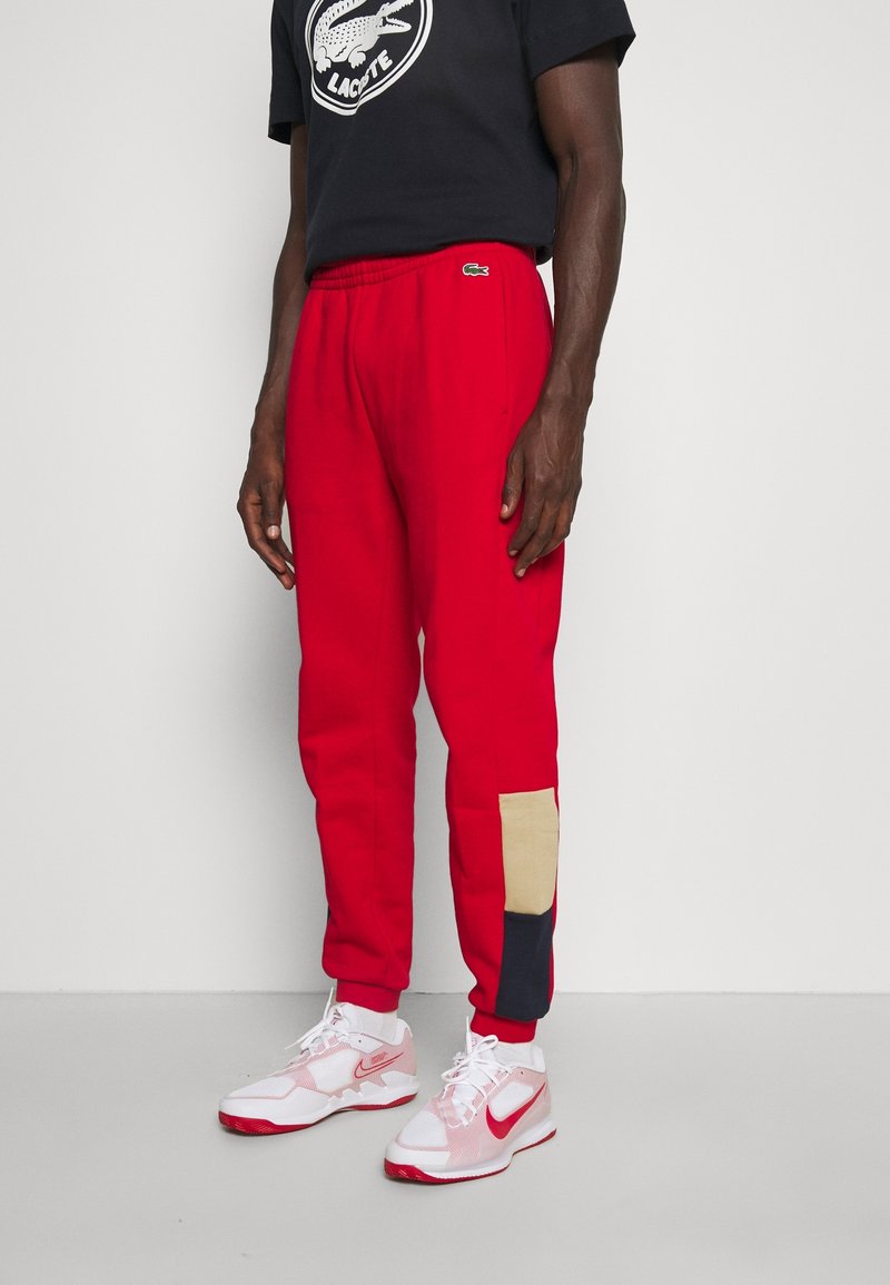 Rote Sweatpants mit einem elastischen Bund, ausgestattet mit einem beigen und marineblauen Patch am linken Bein, kombiniert mit weißen und roten Sportschuhen.
