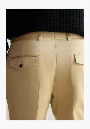 Pantalons kaki avec une texture lisse, dotés de deux poches arrière avec fermetures à bouton et passants de ceinture. Coupe ajustée, design minimaliste.