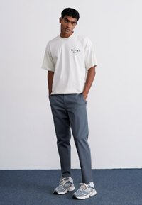 White short-sleeve t-shirt with a black graphic logo, paired with gray slim-fit pants and gray sneakers with white accents.