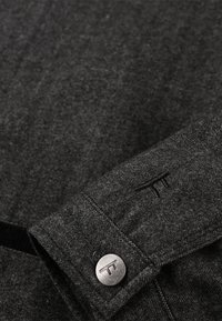 Camisa de tela texturada en gris oscuro que presenta un patrón de espiga, un botón de metal con logo y un detalle cosido en la manga.