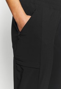 Handen placerad inuti den zipprade sidofickan på svarta byxor med elastisk midja, vilket visar tygtextur och sömnadsdetaljer.