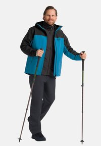 Giacca da hiking blu e nera, con cappuccio, design a strati e chiusura con zip; abbinata a pantaloni scuri e bastoni da trekking.