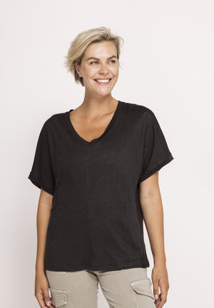 Lächelnde Frau mit kurzen blonden Haaren, die ein lockeres schwarzes V-Ausschnitt-Shirt und hellbeige Cargohosen trägt, vor einem schlichten weißen Hintergrund.