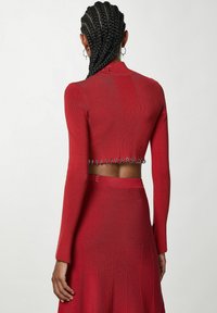 Červený úpletový top s dlouhými rukávy a otevřeným zády, zdobený stříbrnými kovovými prvky. V kombinaci s odpovídající plisovanou sukní.