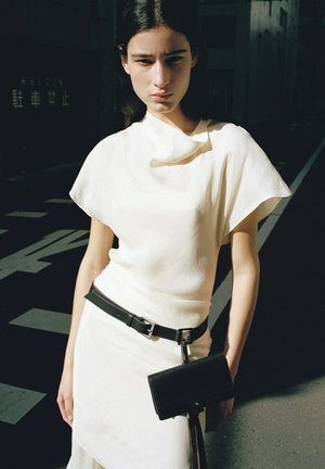 Młoda kobieta stojąca na zewnątrz, ubrana w białą sukienkę z czarnym paskiem, z czarną torebką zawieszoną na pasku, stojąca w świetle słonecznym.