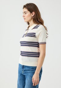 Hellblaues Polohemd mit kurzen Ärmeln in Creme mit navyblauen und roten horizontalen Streifen, das ein strukturiertes Zickzackmuster und einen Kragen aufweist, getragen mit blauen Jeans.
