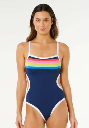 Kvinde iført en navyblå heldragt med hvide kanter og horisontale regnbuestriber på brystet, set forfra.