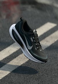 Fekete sportcipő texturált felsőrésszel, fényvisszaverő ezüst swoosh logóval és párnázott fehér talppal. Fűzős és logó részletekkel rendelkezik.