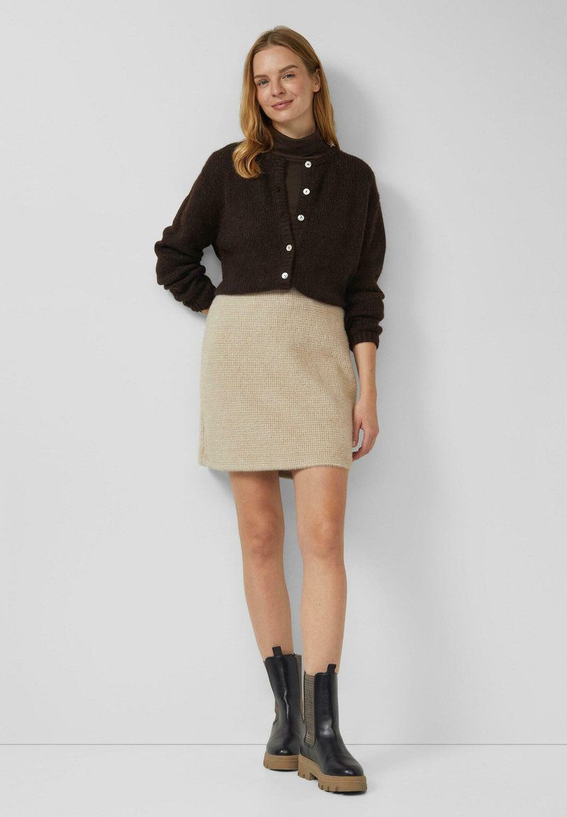 Vrouw draagt een donkerbruine cardigan, beige gebreide rok en zwarte enkellaarsjes, staand tegen een effen lichtgrijze muur.