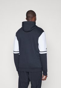 Sweatshirt à capuche bleu marine et blanc avec des manches contrastantes, tissu texturé, et des rayures noires sur les bras et l'ourlet.