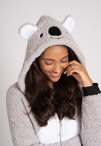 Grijze pluche onesie met een capuchon featuring een koalagezicht. Details omvatten een zwarte neus en witte ooraccenten. Voorkant met rits en een witte bontbuik.