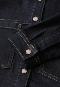 Camicia in denim blu scuro con cuciture gialle a contrasto, colletto appuntito e bottoni metallici con marchio per la chiusura.