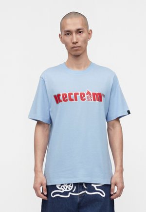 Homme adulte au crâne rasé portant un t-shirt bleu clair avec l'inscription rouge "icecream" et un pantalon foncé avec des motifs graphiques blancs.