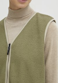 Olive zelena flis brezrokavnik z zadrgo, kontrastnim bež robom, oblečen čez svetlo bež turtleneck pulover, ki prikazuje mehak teksturo.