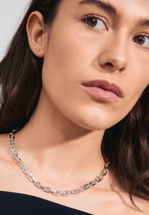 Collier en chaîne argentée avec des maillons rectangulaires à la finition polie, porté autour du cou, complétant un haut sombre.